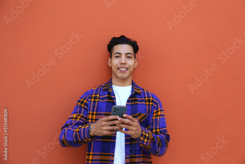Young man of hispanic heritage wearing colorful flannel shirt leaning on the orange wall and typing on his phone. Copy space for text, close up, background.