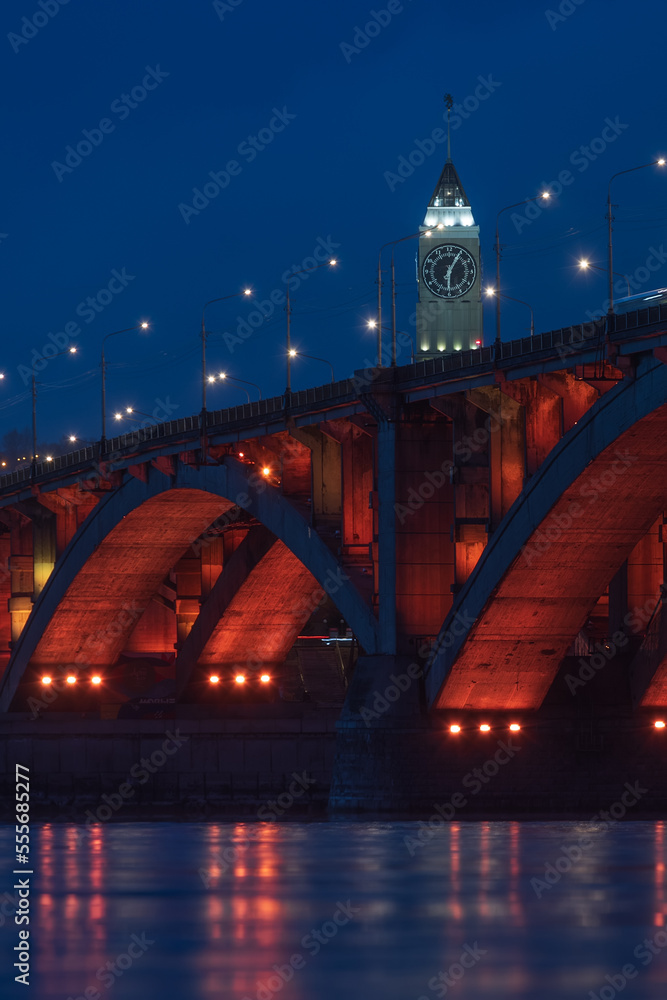 Obraz premium Evening cityscape with red illuminated of Communal Bridge and Clock tower in Krasnoyarsk, Russia. View of the Yenisey River at dusk. Blurred reflection of lighting lights in the rippled water surface