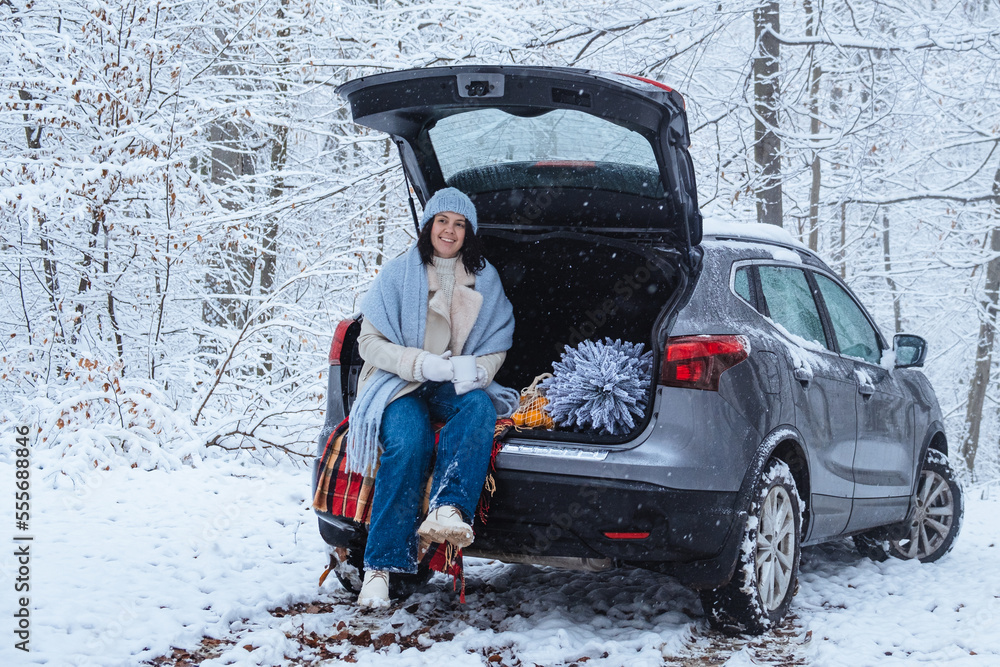 a young woman with a Christmas tree sits in the trunk of a car in the middle of a winter forest