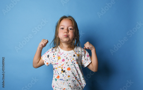 Vászonkép a little girl shows strength by raising her arms up and puffing out her cheeks on an isolated blue background