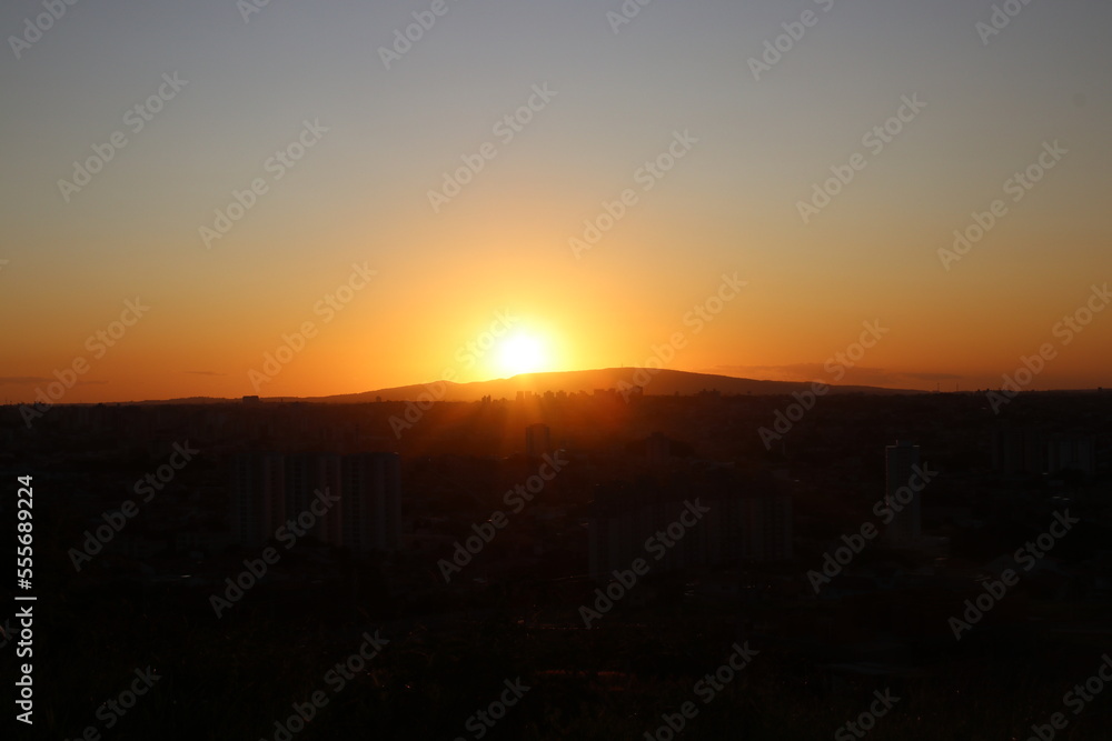 landscape with sunset on the mountain, city, buildings and houses, lights, sunshine and sky in colors yellow, orange and blue