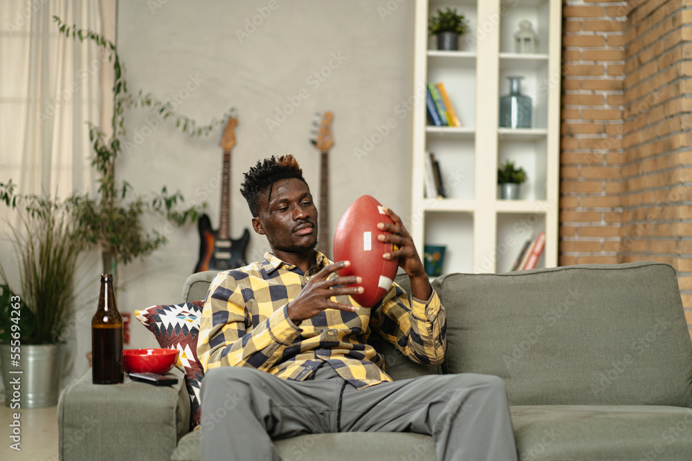 Obraz premium Young African man watching sport match on television and supporting his favorite team. Happy man enjoying victory, watching sports competition on tv