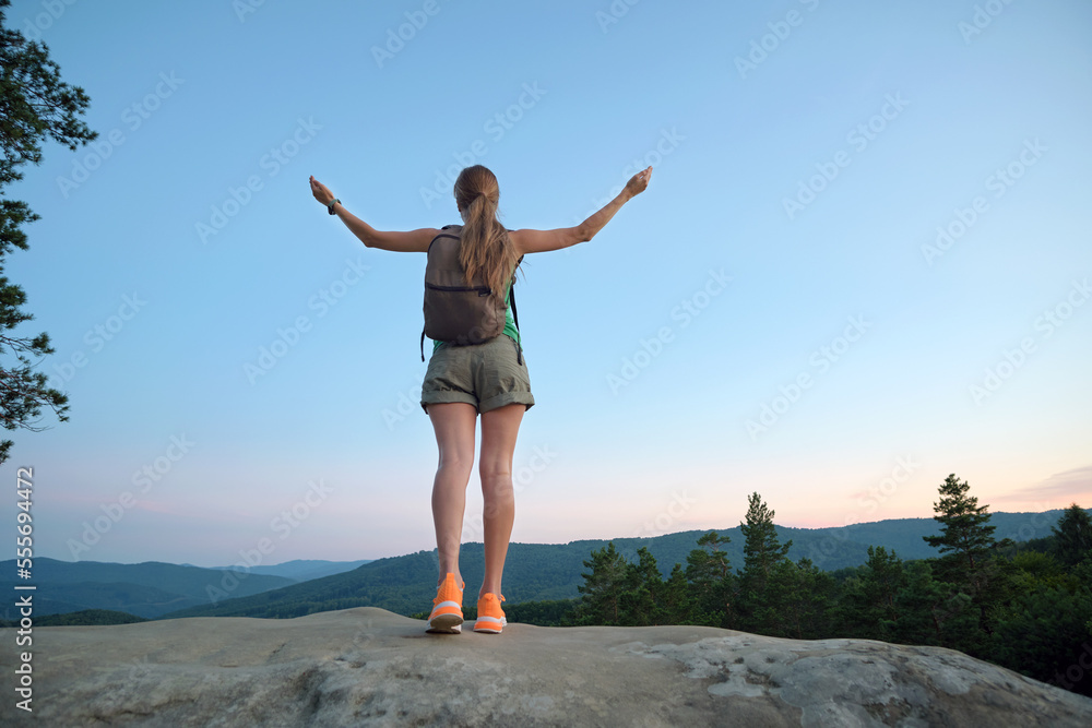 Young woman hiker standing alone with outstretched arms on mountain footpath enjoying view of evening nature on wilderness trail. Active way of life concept Young woman hiker standing alone with outstretched arms on mountain footpath enjoying view of evening nature on wilderness trail. Active way of life concept