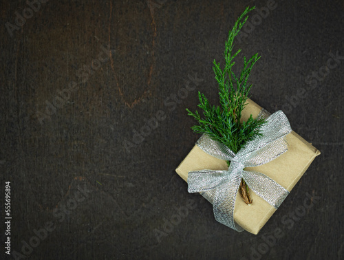 Small gift in kraft paper with a sprig of juniper .Box with silver ribbon lies on a dark brown background .
