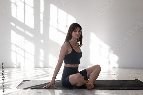 Beautiful fit woman sits on yoga mat before practice stretch exercises in a loft studio with sunbeams on the wall