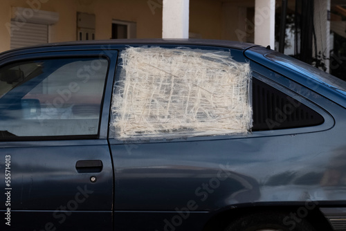 a white car with broken glass on the passenger door wrapped in white film and sealed with white painter's tape. An old wrecked car against a background of green foliage.