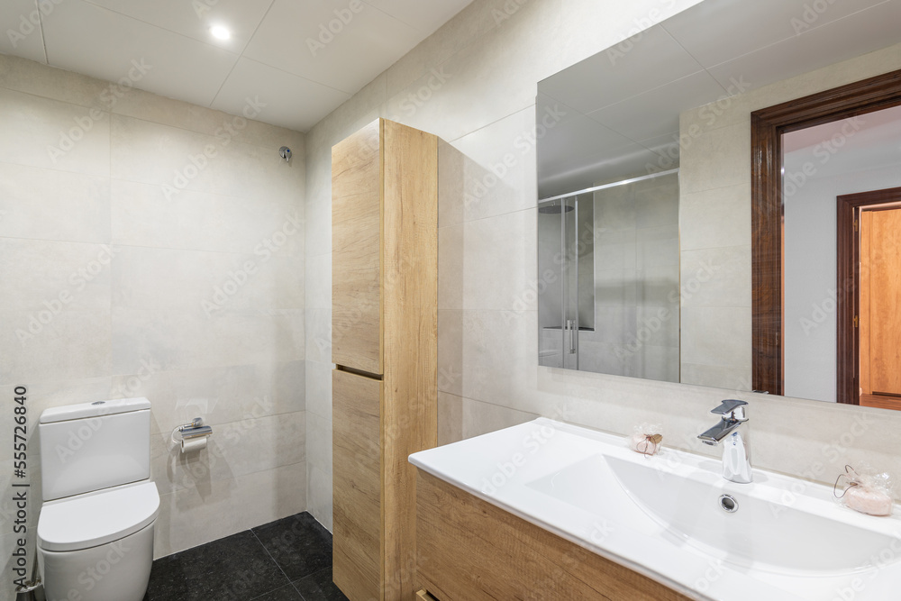 Modern bathroom with white tiled walls. Small wooden cabinet for toiletries and towels sits against wall and harmonizes in color with cabinet for sink. Mirror reflects doorway into room.