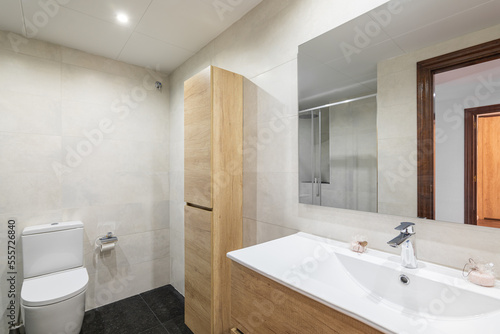 Modern bathroom with white tiled walls. Small wooden cabinet for toiletries and towels sits against wall and harmonizes in color with cabinet for sink. Mirror reflects doorway into room.