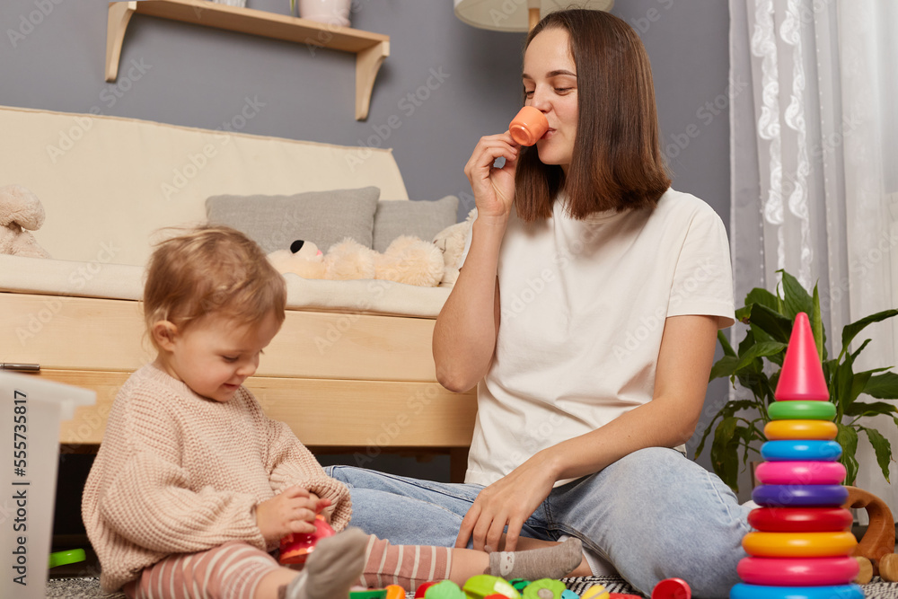 Naklejka premium Happy mother and toddler daughter playing games with toys together in comfortable cozy house, having fun. Little girl and mummy spending leisure time, pretending drinking from toys dish.