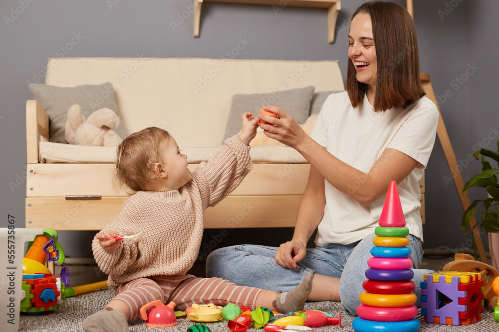 Indoor shot of mom and daughter together sitting on floor near cough and playing with toys, mommy having fun with her toddler kid, expressing positive emotions. Indoor shot of mom and daughter together sitting on floor near cough and playing with toys, mommy having fun with her toddler kid, expressing positive emotions.