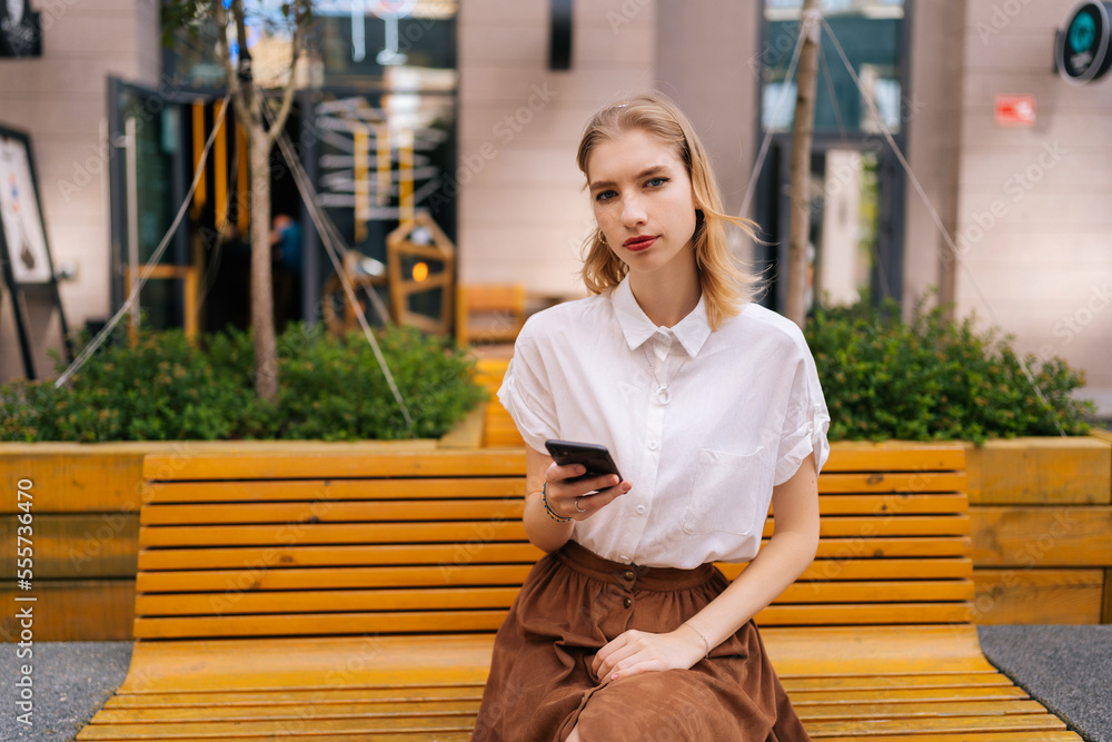 Portrait of confident charming young woman sitting on bench and looking at camera holding mobile phone in hand on background of building. Serious female using mobile phone outdoors in summer. Portrait of confident charming young woman sitting on bench and looking at camera holding mobile phone in hand on background of building. Serious female using mobile phone outdoors in summer.