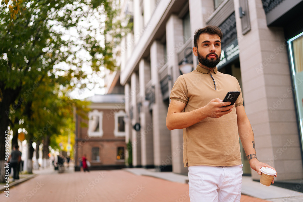 Fototapeta premium Low-angle view of confident young man with tattooed hand walking on city street, using smartphone and drinking takeaway coffee on summer day. Bearded hipster male typing mobile phone.