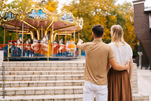 Rear view of unrecognizable loving young couple standing embracing watching rotation of carousel in amusement park. Happy man pointing hand and woman walking spending time together on warm summer day. © dikushin