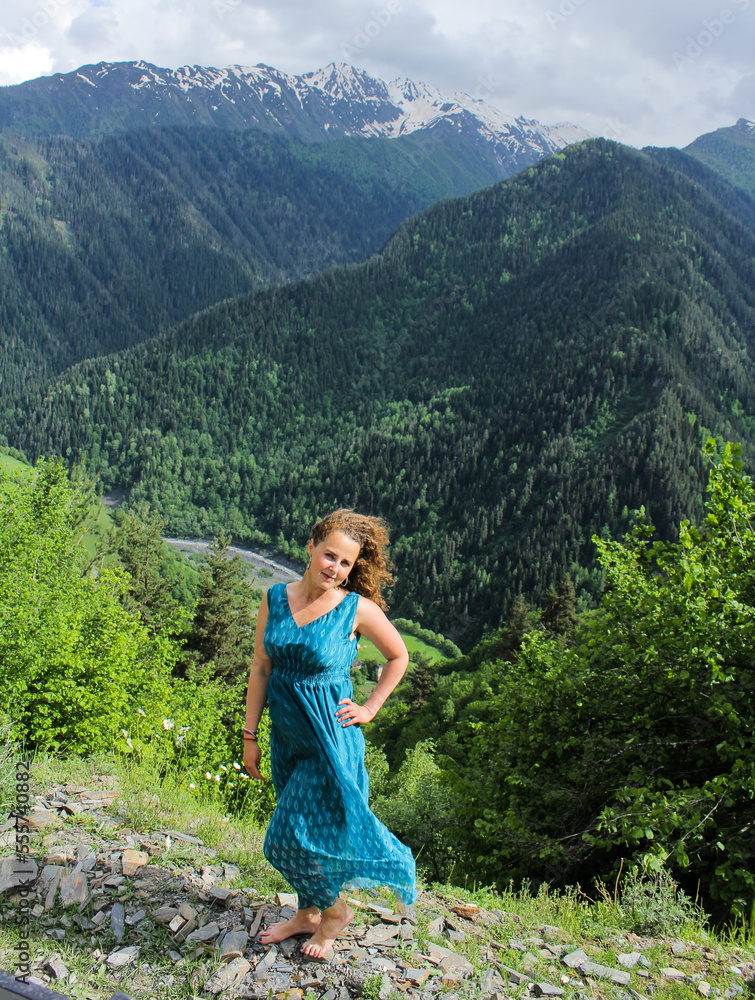 Elegant woman in blue dress at the mountains. Happy girl with wavy hair. Freedom concept. The background of the majestic mountains of the Caucasus and wooden fence. Svaneti, Georgia.