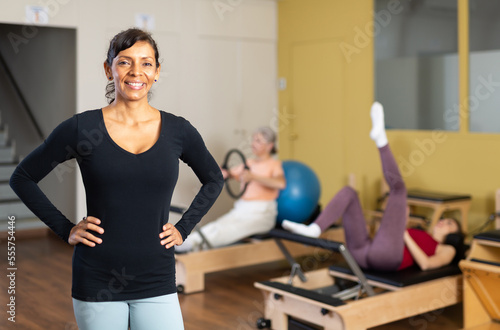 Portrait of sporty smiling Hispanic woman standing with arms akimbo in pilates studio during group workout..