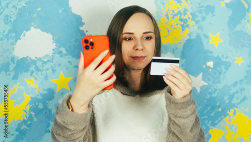 Portrait of beautiful woman in plush hoodie browsing smartphone, entering card details, paying internet purchase on background of colorful bright wall.