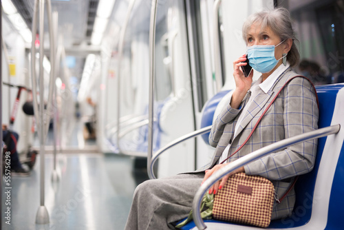 Senior woman in face mask talking on phone while sitting inside subway train.