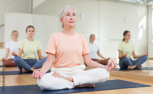 Senior woman sitting on mat in lotus pose during group yoga training with her family.