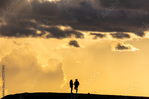 silhouette of a couple, woman and man, standing on a hill. Bright orange sunset sky with high building clouds in the background. 