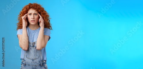 Confused panicking young distressed redhead curly girlfriend frowning touch temples cannot focus look perplexed worried not remember trying pull emotions together standing blue background photo