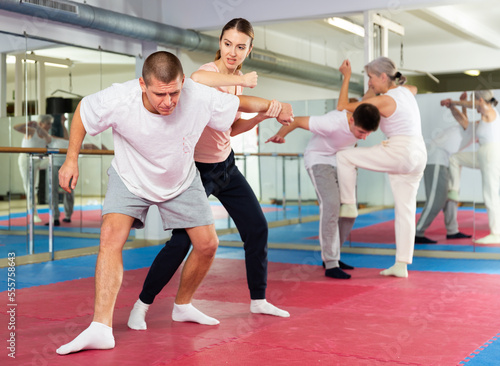 Concentrated sporty young woman learning self defence techniques in sparring with man, practicing elbow blow with wristlock to opponent in gym