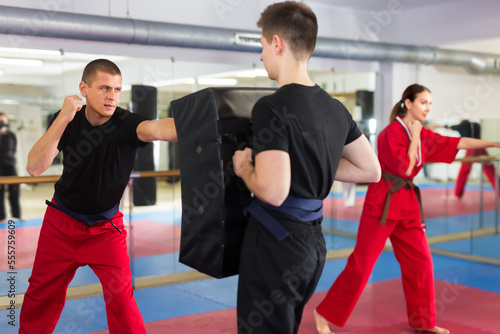 Focused sporty man learning to punch in martial arts training, exercising on kick shield held by young assistant in gym