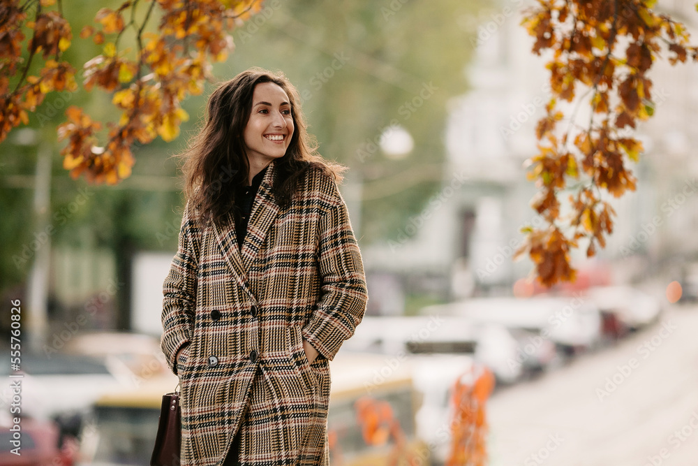 Side view of happy girl with dark hair, dressed in plaid coat and holding hands in pocket while walking on city street during autumn weather and looking away Side view of happy girl with dark hair, dressed in plaid coat and holding hands in pocket while walking on city street during autumn weather and looking away