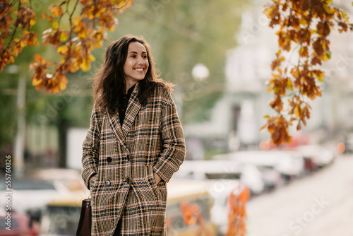 Side view of happy girl with dark hair, dressed in plaid coat and holding hands in pocket while walking on city street during autumn weather and looking away