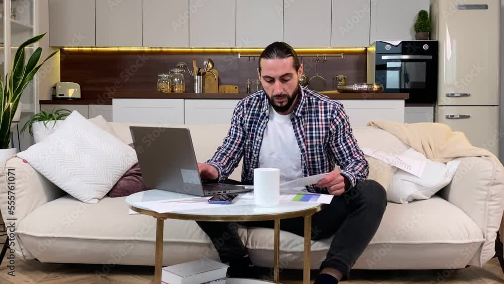 Middle aged bearded man working at home by using laptop. Positive male worker typing, checking paper graph chart while using computer indoors. Cheerful manager having remote job via Internet Middle aged bearded man working at home by using laptop. Positive male worker typing, checking paper graph chart while using computer indoors. Cheerful manager having remote job via Internet