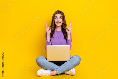 Full body photo of cheerful lady sit floor use netbook look indicate fingers up empty space isolated on yellow color background © deagreez