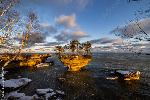 Sunrise illuminates this sculpted formation known as Turnip Rock on the Lake Huron shoreline in early winter. Pancake ice can be seen floating in the surrounding waters. Blue sky, puffy clouds.