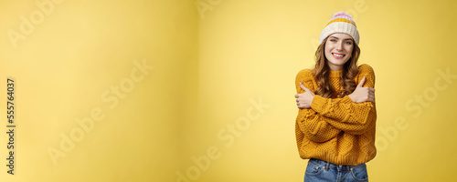 Getting cold bring jacket. Portrait charming tender feminine cute girlfriend embacing herself cuddling smiling warm gaze freezing outside standing knitted sweater hat, yellow background photo