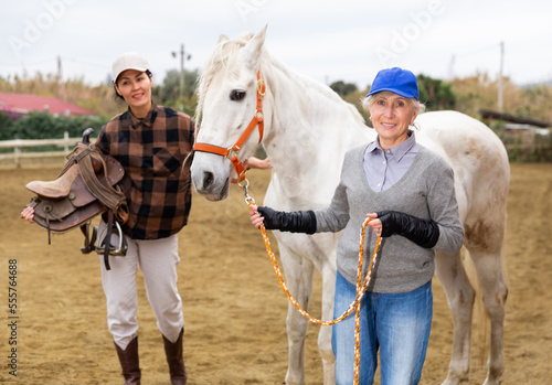 Women ranchers preparing white horse for ride. Senior European woman leading horse, Asian woman carrying saddle.