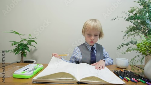 Cute 5 years old caucasian boy dressed very smart does homework in a big notebook drawing with pencil. Rulers, alidad and colorful pencils are also on the table