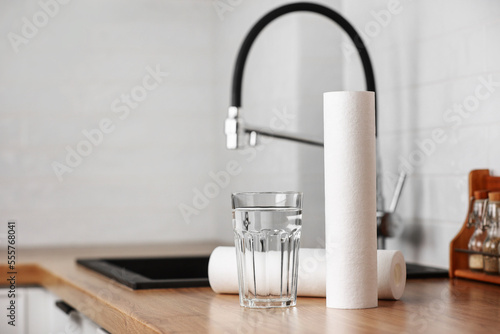 A glass of clean water and foamed polypropylene filter cartridges on wooden table in a kitchen interior. Installation of reverse osmosis water purification system. Concept Household filtration system.