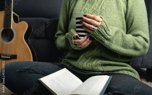 Unrecognizable woman with cozy knit sweater holding cup of coffee and reading book in the living room carpet