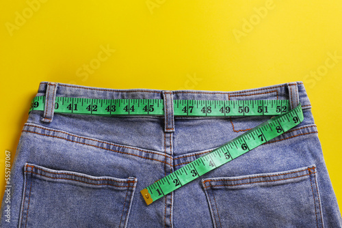 Jeans with measuring tape on yellow background, top view. Weight loss concept