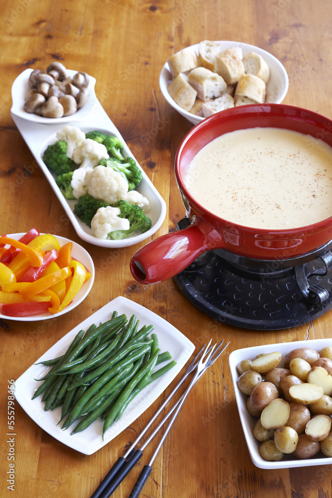 Cheese fondue in red pot with dishes of vegetables and bread on a wooden table Cheese fondue in red pot with dishes of vegetables and bread on a wooden table