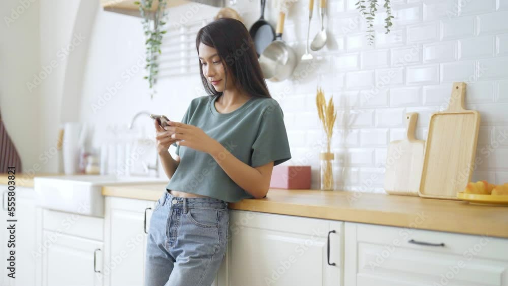 Happy young asian woman relaxing at home. Asia female standing at counter kitchen and using mobile smartphone Happy young asian woman relaxing at home. Asia female standing at counter kitchen and using mobile smartphone