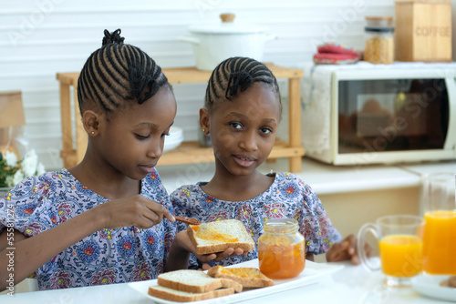 Twin African girls making breakfast , spreading jam on slices of toast and eating in modern kitchen, eat delicious together