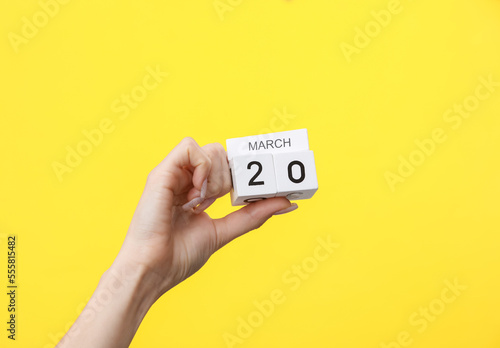 Woman's hand holding wooden block calendar with the date march 20 on yellow background