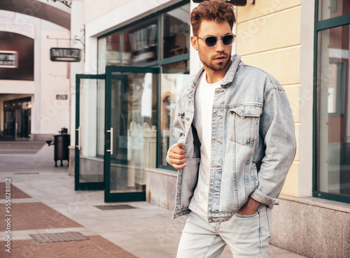 Portrait of handsome confident stylish hipster model. Sexy man dressed in jeans jacket. Fashion male posing in the street. In sunglasses in Europe city. Outdoors at sunset. .in the alley of boutiques © halayalex