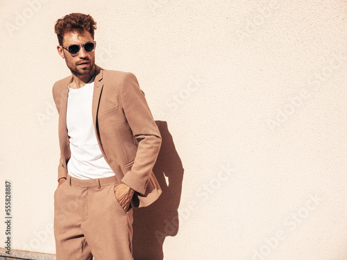 Handsome confident stylish hipster lambersexual model.Sexy modern man dressed in elegant beige suit. Fashion male posing in the street in city at sunset. In sunglasses. Near wall