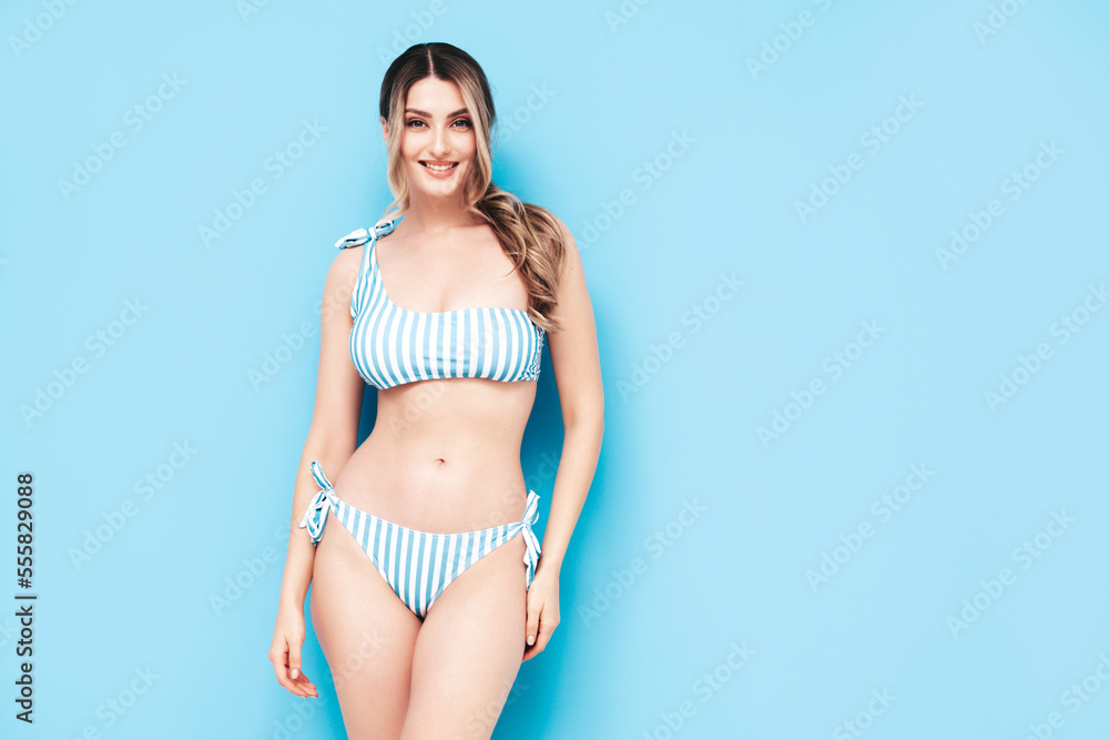 Fototapeta premium Young blond model in summer striped swimwear bathing suit. Sexy carefree woman having fun and going crazy. Female posing near blue wall in studio. Cheerful and happy. Perfect body