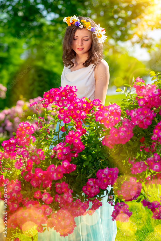 Naklejka premium Spring sexy fantasy woman enjoys standing near flowering pink roses. Outdoor portrait of young beautiful happy smiling lady posing. Youth, romantic and lifestyle concept. Copy, empty space for text