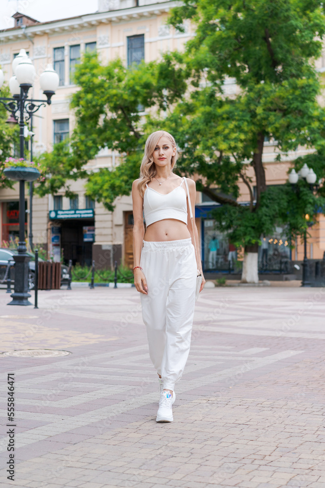 Young beautiful stylish woman walking in white tracksuit, holding handbag in her hands, smiling happy, street style, spring summer trend, caucasian blonde woman outdoors in the city