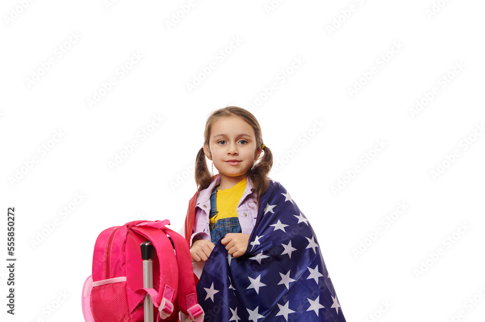 Charming little girl, a smart school kid, wrapping in USA flag, standing near a backpack, looking at camera, on white background. Concept of studying abroad. English courses. British American schools Charming little girl, a smart school kid, wrapping in USA flag, standing near a backpack, looking at camera, on white background. Concept of studying abroad. English courses. British American schools