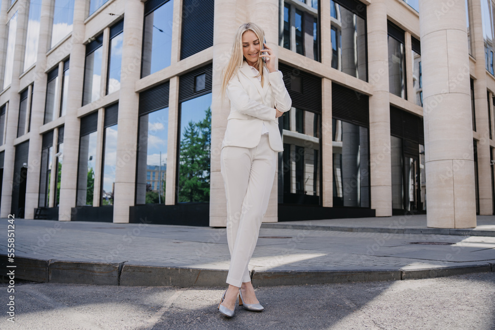 Pretty Italian businesswoman in white suit standing outside talking by phone, toothy smiling. Cheerful student received good news. Gorgeous Swedish female satisfied by her career. Business, education.