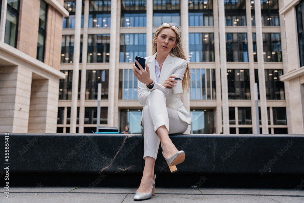 Tired blonde caucasian businesswoman sitting against blurry building thinking, looking aside. Italian pretty student girl waiting for exam results outdoors. Female entrepreneur having coffee break.