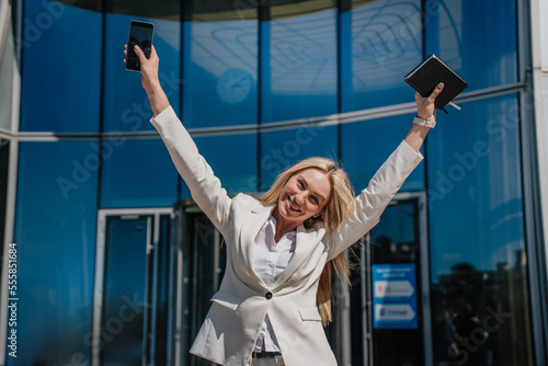 Independent young blonde Italian businesswoman in white suit spread hands in a winner expression holding diary and phone over business center. Successful lawyer celebrates win. Business and finance.